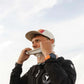Person wearing a cap and jacket with a logo, adjusting their cap against a cloudy sky.