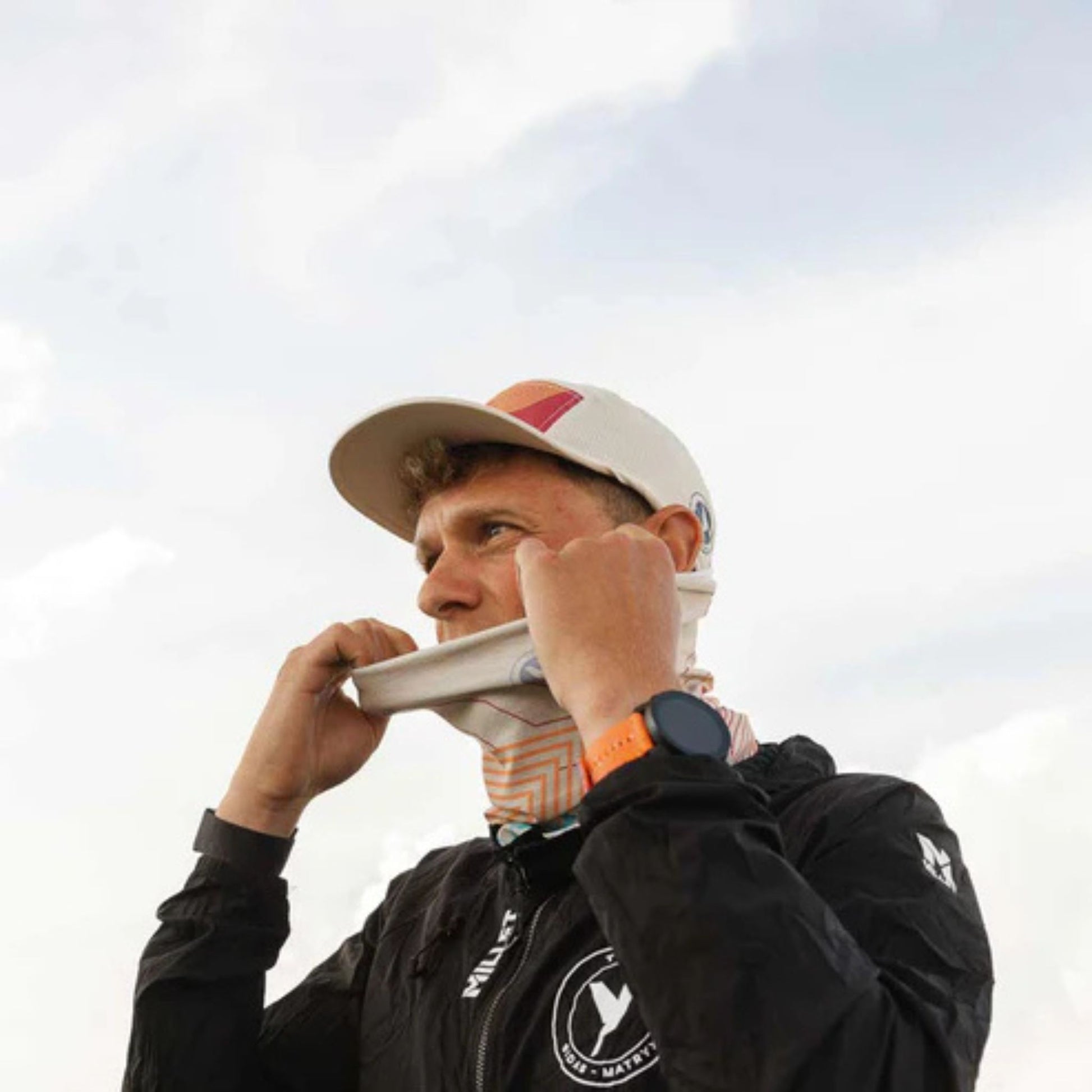 Person wearing a cap and jacket with a logo, adjusting their cap against a cloudy sky.